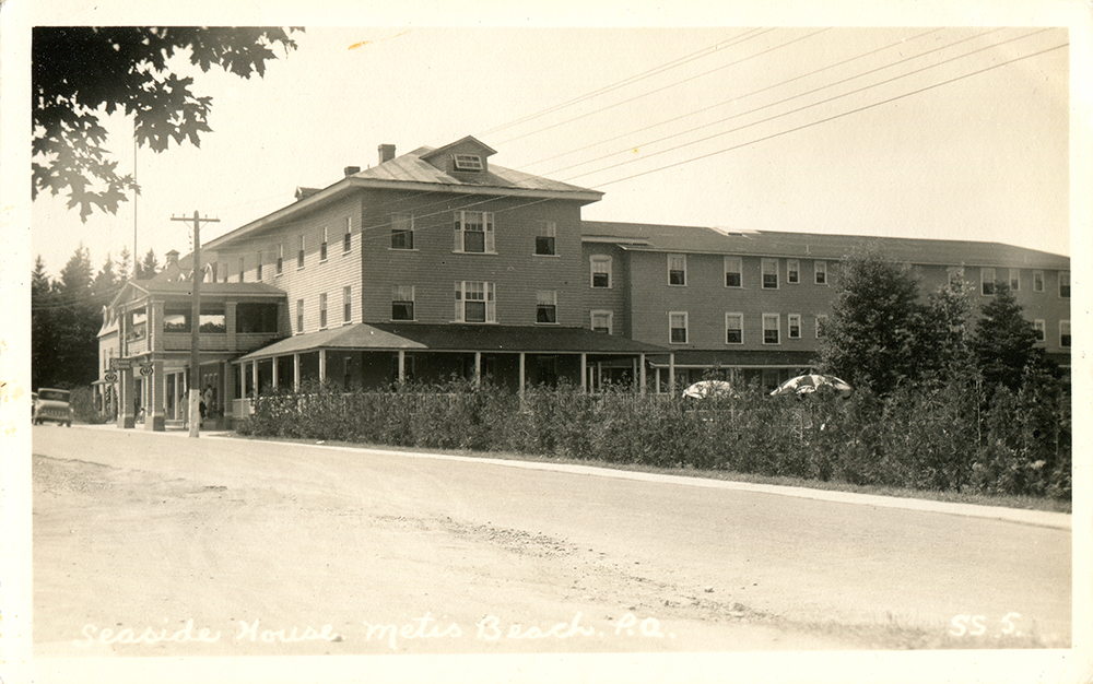 Postcard presenting a black and white photograph of the Seaside Hotel of Metis Beach. The imposing three-storey wooden hotel has two wings. One runs along the road and the other runs backwards from the center of the first wing. The courtyard is hidden by a young cedar hedge , where that reveals two sun umbrellas are seen.