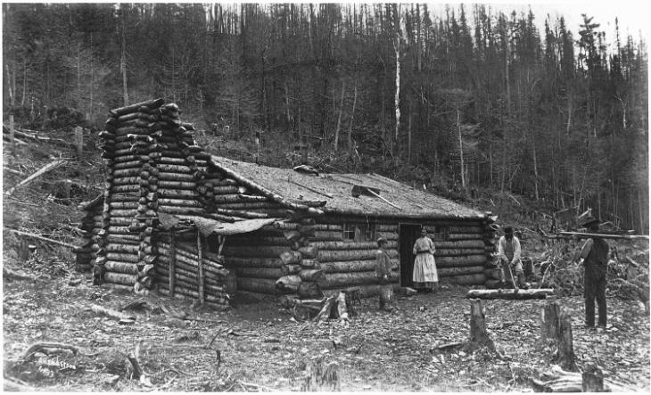 Black and white photograph of a rustic log house. The house is located at the foot of a hill and is surrounded by many tree stumps of the trees that were probably used to build the house. Four people are outside the house. A woman in a white dress is standing in the doorway and looking at us. A few steps away from her stands a young boy holding a wooden bucket. He observes a man working to remove the bark from a section of tree on the ground. A man approaches the scene carrying a load on his shoulders.