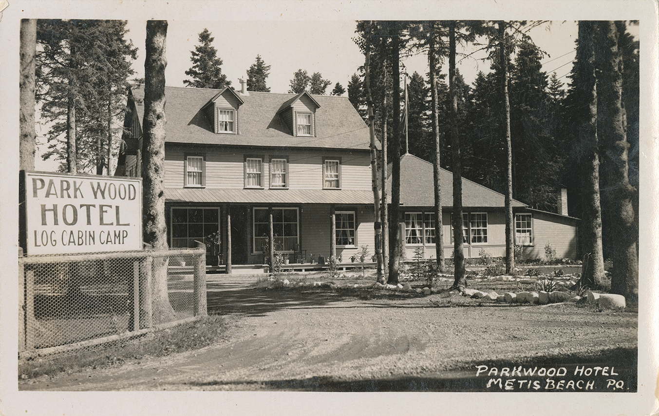 Postcard showing a black and white photograph of the entrance of the Park Wood Hotel. The hotel is a large three-story house with a one-story annex. The hotel is made entirely of wood and is surrounded by a spruce forest. In the foreground of the photograph is the access road to the hotel. On a wire mesh fence is the hotel's wooden sign, which says Park Wood Hotel Log Cabin Camp.