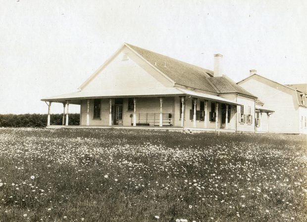 Black and white photograph of the north and west facades of Estevan lodge. The exterior of the building is in wood and the gable roof is made of cedar shingles. The windows are equipped with shutters. The fishing camp is equipped with a generous verandah on which is placed rack for fishing rods. At the back of the main building are outbuildings occupied by the owner's servants. The two-storey building is slightly set back to the west in relation to the main building. The building is surrounded by a flowering meadow. We can see the huge cedar hedge that occupies the front space of the fishing camp. In spite of its size, the fishing camp is rather sober, only the columns of the verandah roof underline the Victorian style of the building's architecture.