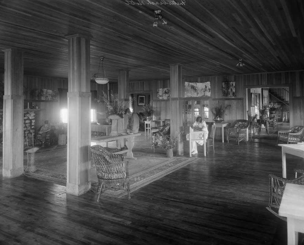 Black and white photograph of the interior of the vast lobby of the Boule Rock Hotel. Entirely made of wood, the lobby is bright and tidy. In the center of the space, a carpet covers the wooden floor and we can see a portion of the fireplace mantle made of field stones. Rattan armchairs and tables are distributed throughout the room. Bouquets of flowers are also found on most of the tables and on the floor. Two men and one woman are also seen. The woman is sitting at a desk and seems to be writing a letter. One of the men is sitting at a console table and appears to be reading. The last man sits in an armchair by the fireplace reading his newspaper.