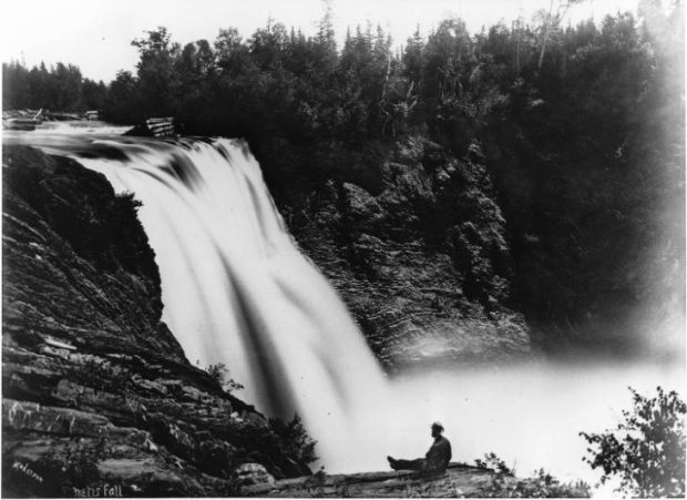 Black and white photograph of the fall on the Metis River. The 90 foot waterfall is bordered by two steep rocky cliffs surrounded by forest. The photographer captured the waterfall from the top of the cliff on the left side. A few meters from where the photograph was taken, a man is sitting with his legs stretched out on the edge of the cliff. He is watching the waterfall and its rapid current. The photographer has made a long exposure, the river water flows from the top of the waterfall in a large white stream.