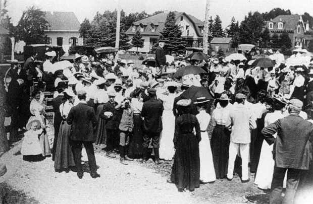 This is a black and white photograph of a gathering of people at the celebration of Queen Victoria's Jubilee in 1897. People are gathered outside in the midday sun. In the center of the scene, a man stands overlooking the crowd around him. In the background are some of the stately homes of Metis Beach.