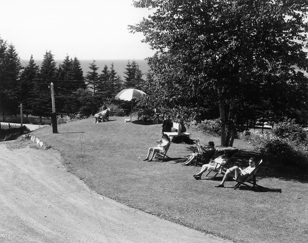 Black and white photograph of vacationers sitting in lawn chairs. Some are in the sun, others are in the shade under a tree or an umbrella. They are located on a landscaped area. In front of them passes a dirt road, possibly the driveway to their hotel. In the background past spruce trees is the St. Lawrence River.