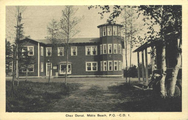 Black and white photograph of the Chez Donat hotel. The two-storey hotel is located on the side of the road. A three-storey turret forms the right corner of the building. Each window is decorated with white curtains. The photograph was taken from the courtyard entrance in front of the hotel.