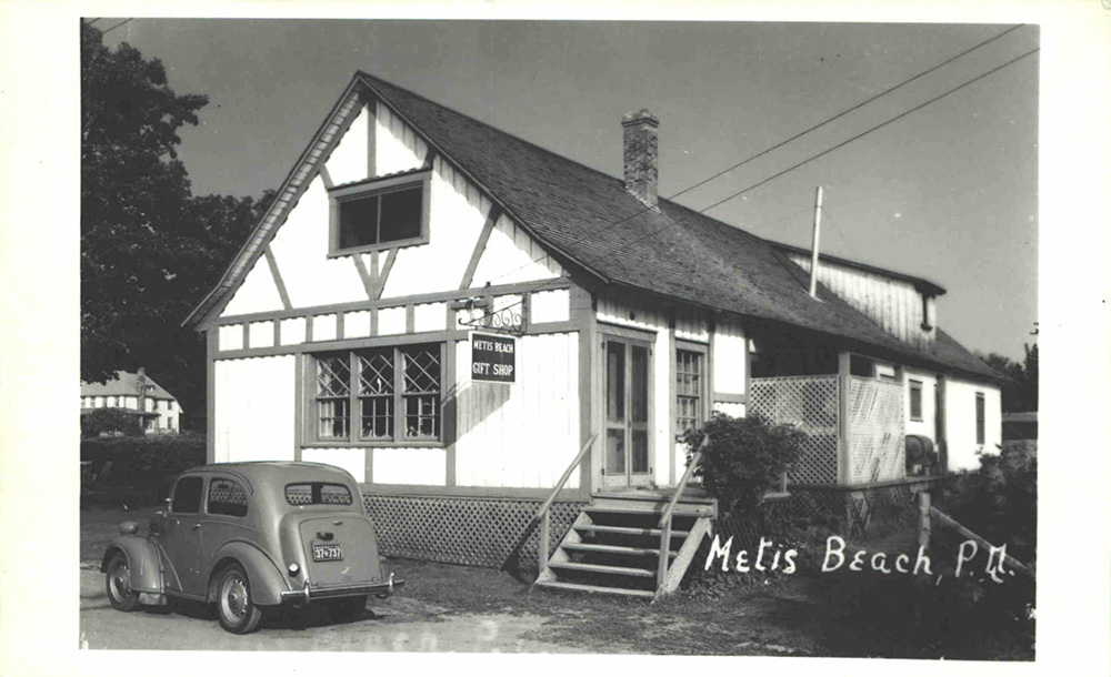 Black and white photograph of the north and west facade of the Metis Beach Gift Shop. The gift store is a white two-storey residence with a cedar shingle roof. A car is parked under the window on the north façade. On this same façade hangs the store's sign, METIS BEACH GIFT SHOP.