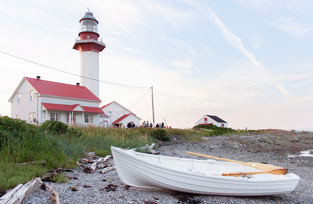 Recent colour photograph of the lighthouse at the end of a summer's day. In the foreground a white rowboat rests on the shore. In the background, we see the keeper's house, the lighthouse, the foghorn building and a small shed. All the buildings are white with red roofs. A small crowd is gathered near the buildings.