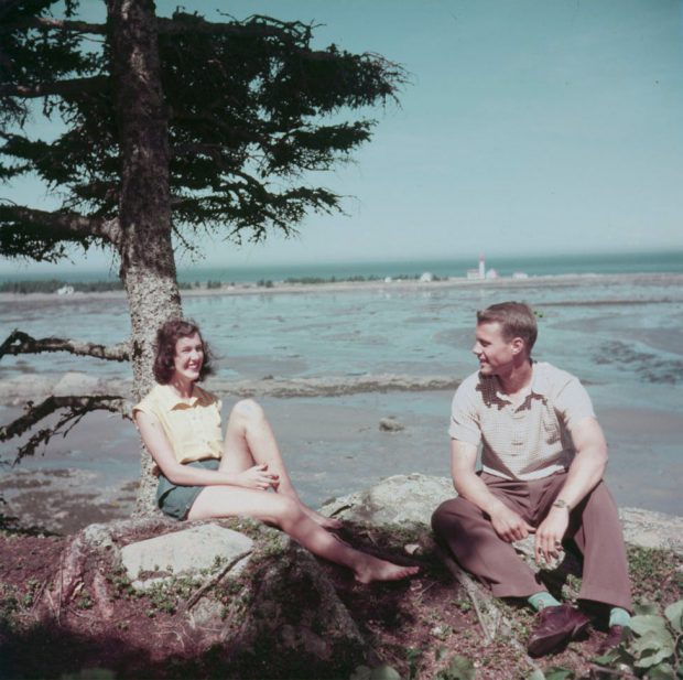 Colour photograph of a young man and woman sitting at the foot of a spruce tree on rocky ground. In the background the St. Lawrence River and the bay at low tide. The lighthouse can be seen at the end of a rocky peninsula.
