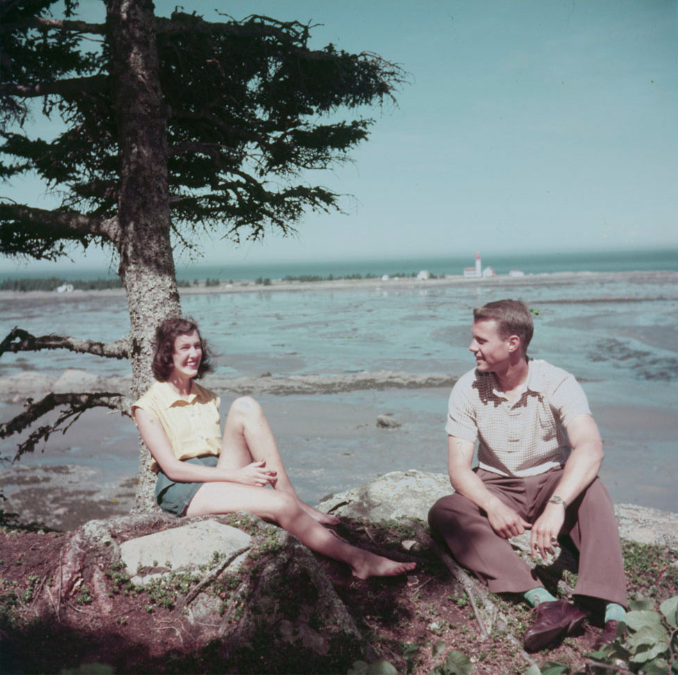Colour photograph of a young man and woman sitting at the foot of a spruce tree on rocky ground. In the background the St. Lawrence River and the bay at low tide. The lighthouse can be seen at the end of a rocky peninsula.