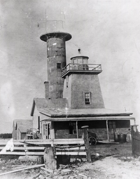 Black and white photograph of the front of the first wooden lighthouse. Behind the first lighthouse appears the cement tower of the new lighthouse which is under construction. Missing is the cast iron and glass top that will house the lantern. This portion has been sketched by hand with a lead pencil on the photograph.