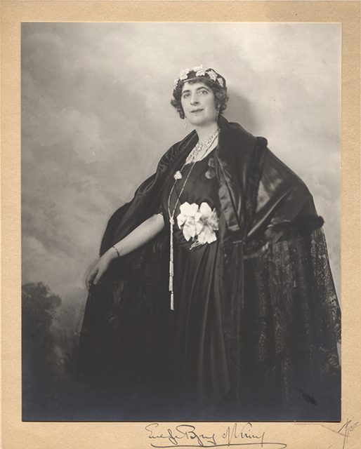 Black and white studio portrait of Lady Byng. Lady Byng poses in front of a painted background of clouds and trees. She is wearing a black cape of satin, lace and fur. A black dress and two white flowers in fabric at the belt. She wears a crown made of maple leaves. Her left hand is resting on her hip allowing to see the details of her cape and dress. Her right arm is outstretched and she is holding her cloak to leave it open. The photograph is mounted on a card signed by her hand "Evelyn Byng of Vimy" and the photographer "Rice".