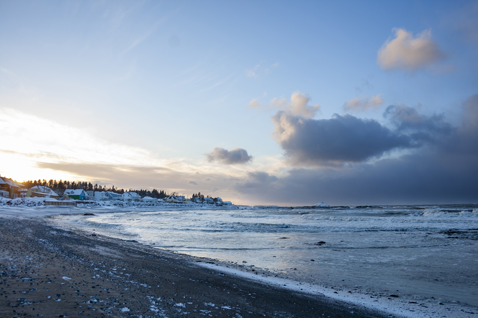 Colour photograph of the shoreline of Metis during winter. Taken from the shore of the St. Lawrence River. The right portion of the photograph shows the river at high tide. The left portion of the photograph shows the residences along the shoreline. The photograph was taken in the late afternoon. A thin layer of snow covers the roofs and the beach.