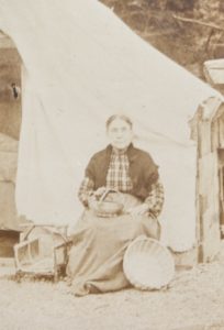A sepia coloured picture of an Indigenous woman seated in front of a tent selling woven baskets.