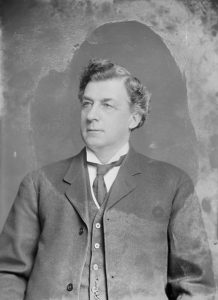 Black and white photograph of Senator John Gillanders Turriff taken while seated and from the waist up. Figure is wearing a period suit and tie.