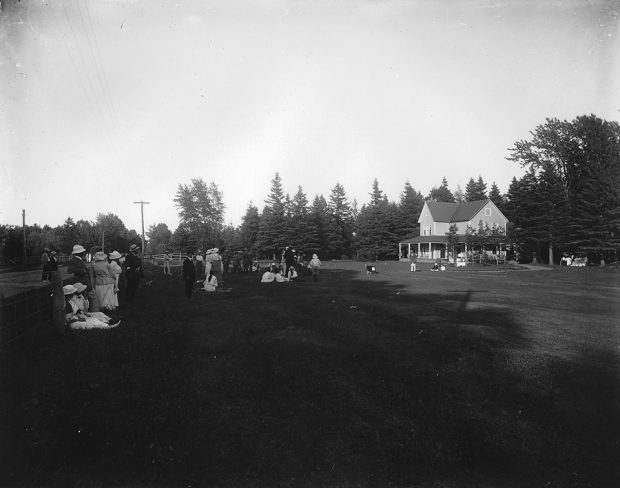 Black and white photograph of the first tee of the Cascade Golf Club. Twenty individuals dressed in the fashion of 1914 watch and wait. Some women are sitting on the grass. In the background, a two-storey cottage with clean Victorian architecture.