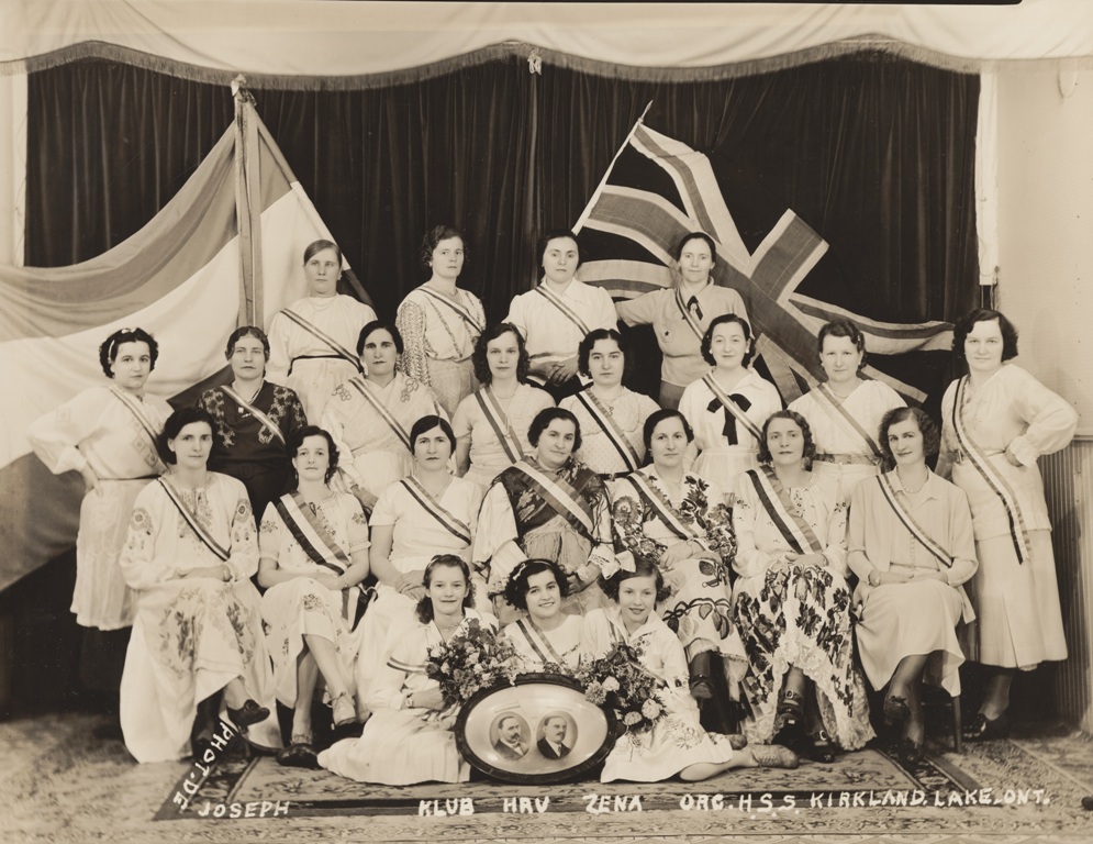 Portrait photograph of four rows of girls and women wearing light coloured dresses and sashes, in front of two flags – the Croatian flag and the Union Jack.