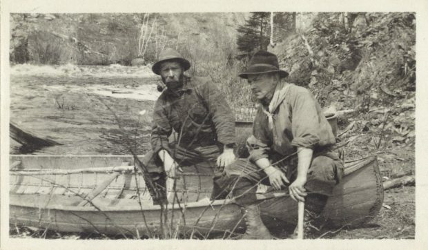 Prospectors are sitting on the edge of their canoe on shore. In the background are river rapids and a tree lined rocky landscape.