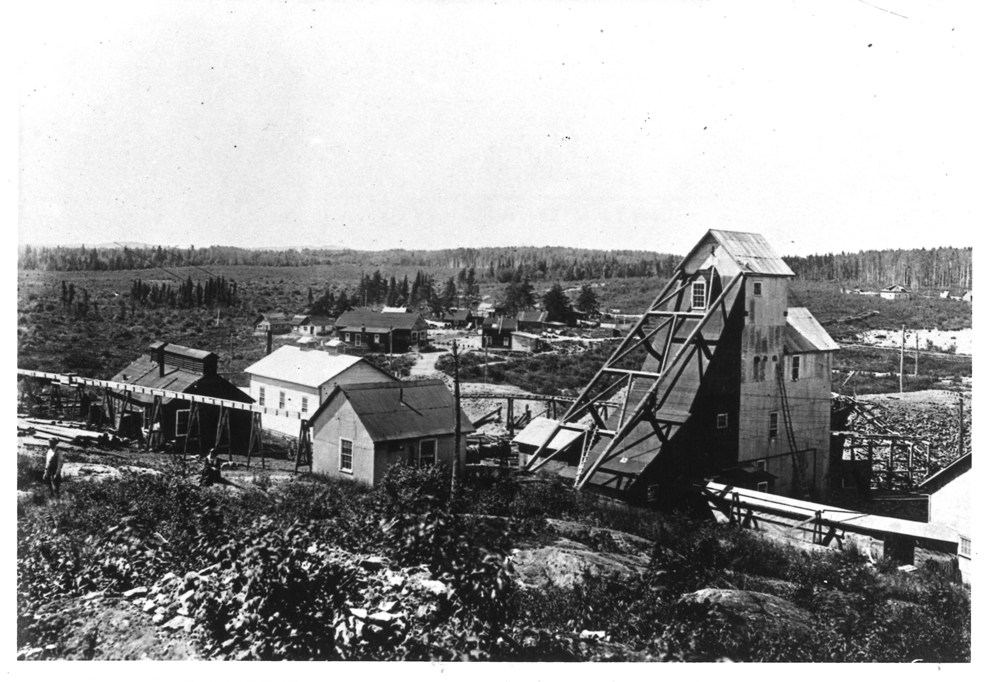 Black and white photograph of the tall headframe of the Tough-Oakes Mine is seen in the foreground with other mine buildings. Small houses line a road winding in a large clearing. Deep forest in the background.