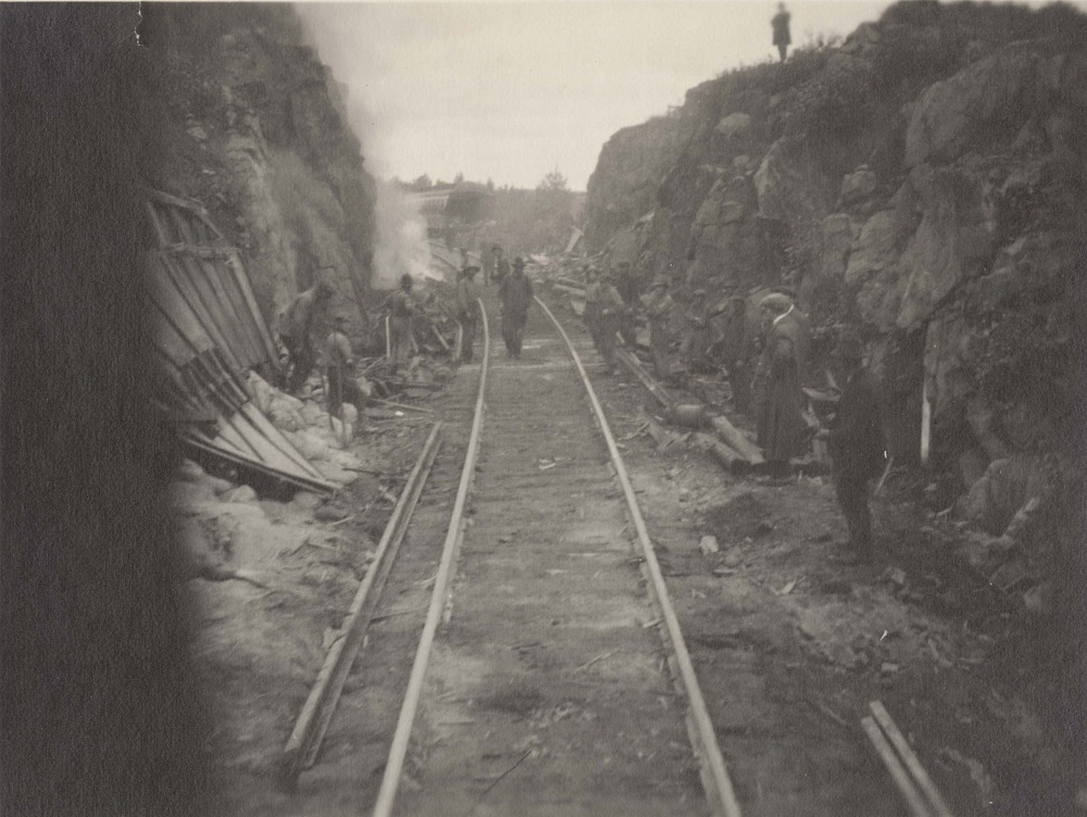 Black and white photograph of the construction of the Temiskaming and Northern Ontario Railway. The railroad tracks lie on flat ground between the blasted rock of the Cambrian Shield. The railroad gang stand in the background.