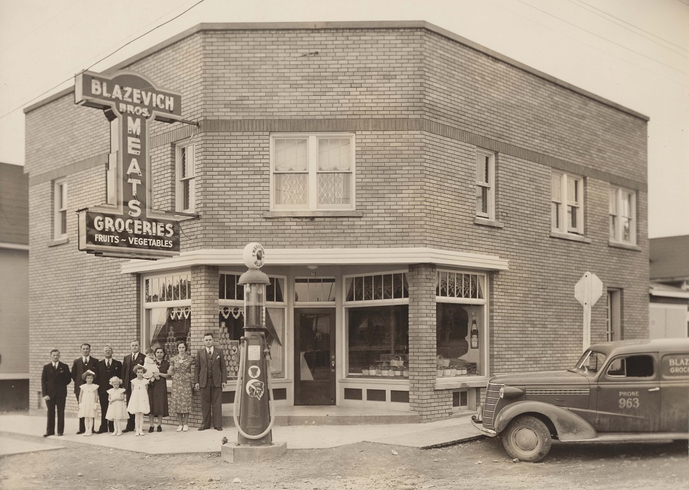 Black and white photograph of the Blazevich Brothers grocery store on Main Street. The Blazevich family is standing underneath the neon sign attached to the brick building. An old fashioned gas pump is in the foreground, and the Blazevich Brothers delivery vehicle is at the right side of the image.