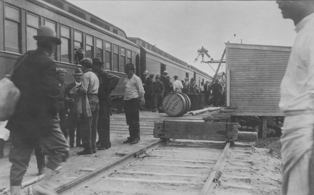Black and white photograph of a passenger train at the Swastika rail station. People are either getting on or off the train.