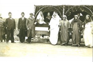 Black and white photograph of people dressed in traditional Syrian clothing with a Coronation Day parade float. Three women and a man are seated on the float, while four men stand in front of the float. Three men in suits and a boy stand to the left of the float, with one leaning against it.