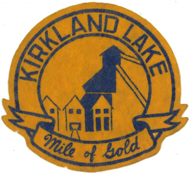 A gold and blue coloured felt patch, with the words Kirkland Lake and Mile of Gold written around the outside of the patch. The image in the centre of the patch is of mine buildings and a headframe in the centre.