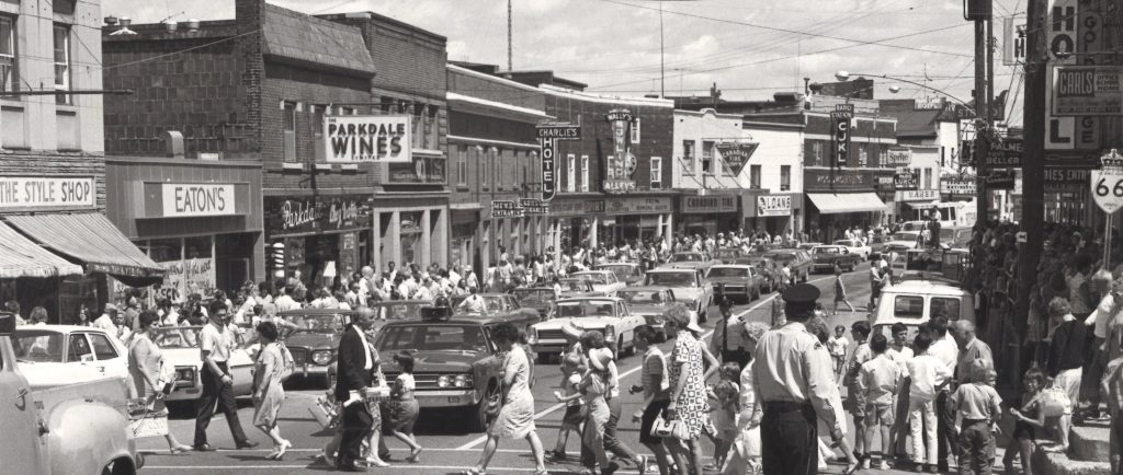 Black and white photograph of a busy street. A policeman directs traffic in the foreground while many people cross the street. Cars line the street, businesses are in the background. A banner hangs over the street.