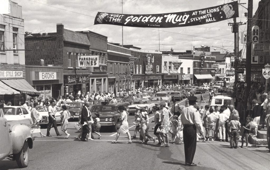 Black and white photograph of a busy street. A policeman directs traffic in the foreground while many people cross the street. Cars line the street, businesses are in the background. A banner hangs over the street.