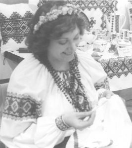 Black and white photograph of a young woman dressed in traditional Ukrainian clothing and doing embroidery on a piece of cloth.