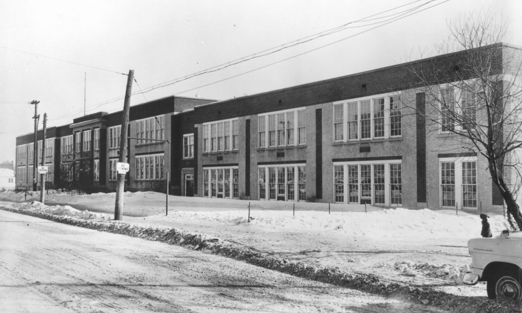 Black and white photograph of a high school, a two-storey brick building with many windows. Snow is on the front lawn. Two telephone poles are at left side of picture, the front end of a white car is in the lower right corner.