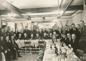 Black and white photograph of Hotelmen’s Banquet in Kirkland Lake Hotel, Charlie Chow is seated, 2nd from left. Over 40 men in suits are seated and standing around tables after a dinner, facing the camera. The tabletops are full of teacups and beer bottles, napkins and empty plates. Four chandeliers hang from the ceiling.