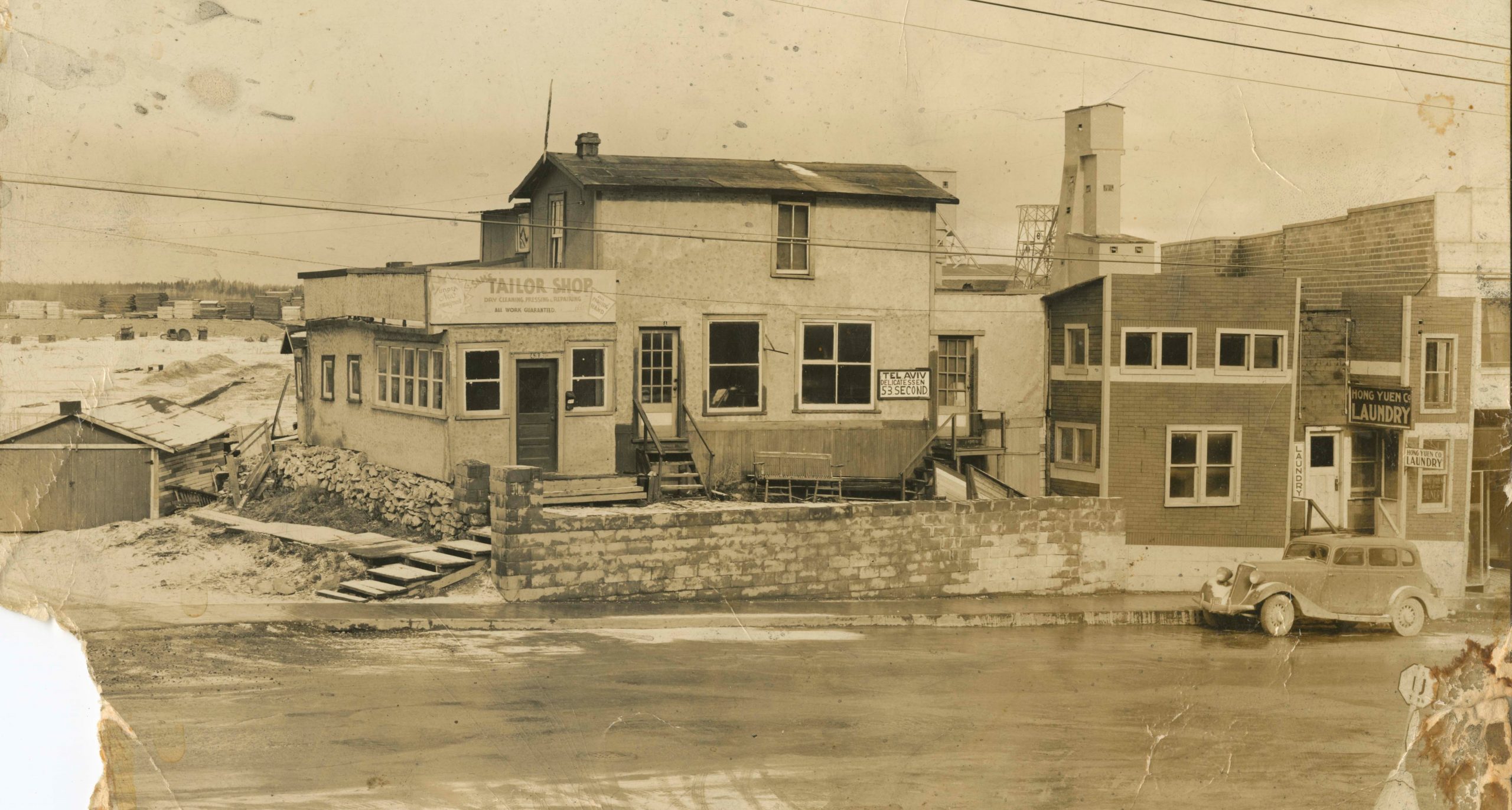 Black and white photograph of a home and businesses on a street with a car parked at the right side of the image. Mine buildings are in the background.