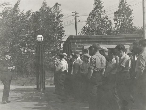 Black and white photograph of a group of men standing beside a large metal gatepost, facing a guard in uniform at the mine property entrance. The guard is holding a piece of paper.