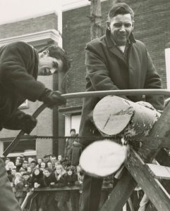 Black and white photograph of a log sawing competition. The man on the left is wearing a dark coat and gloves using a buck saw to slice through a log. The man beside him is also wearing dark clothing and holds the log in place on the rack. A crowd of mostly young people stand behind a barricade in the background, in front of two brick buildings.