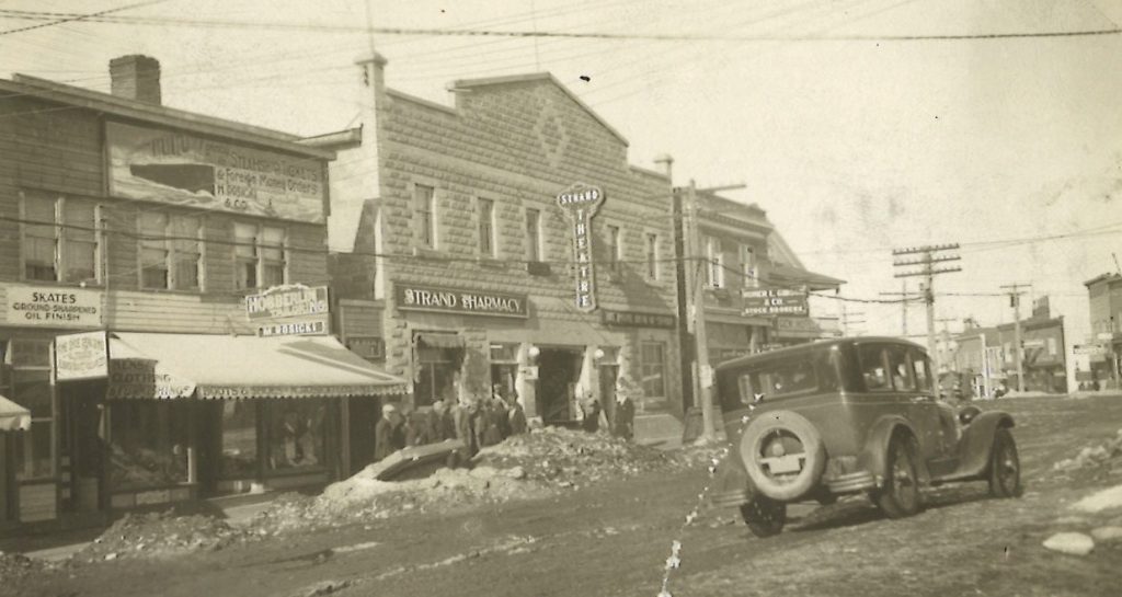 Black and white photograph of a busy street, with cars parked along either side. In the background are several businesses and people standing on the sidewalk. The streets have large snowbanks.