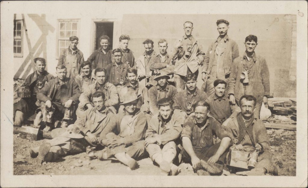 Black and white photograph of 22 miners wearing dirty work clothes of overalls, shirts, jackets, helmets or hats. Some men wear a lantern around their neck, and other men are holding lunch boxes. Men are sitting and standing in the background, in front of a mine building.