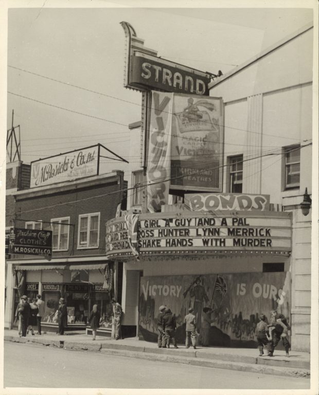 Black and white photograph of the front of the Strand Theatre. The building is white with a large marquee extending over the sidewalk in front. Movie poster and Victory Bond banners hang on the front of the building. Men, women and children are on the sidewalk. A red brick building of storefronts is to the left of the theatre.