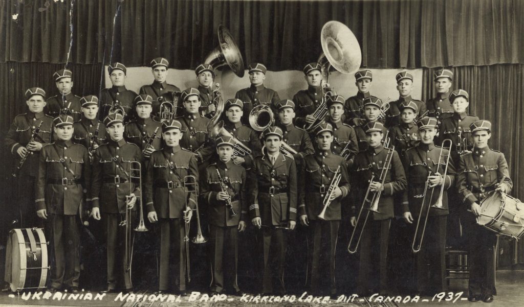 Black and white photograph of three rows of men in uniforms holding instruments in a photography studio. Written along the bottom are the words - Ukrainian National Band - Kirkland Lake, Ont – Canada – 1937 -