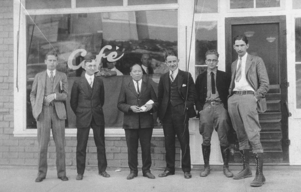 Black and white photograph of six men standing outside of a building with Cafe written on the front window. The men are dressed in suits on the sidewalk.