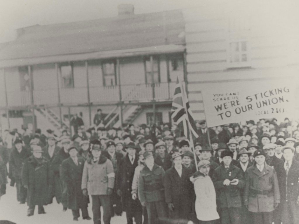 Black and white photograph of a large crowd of men dressed in warm clothing standing outside of a large building in the winter, holding a British flag and a pro-union sign.
