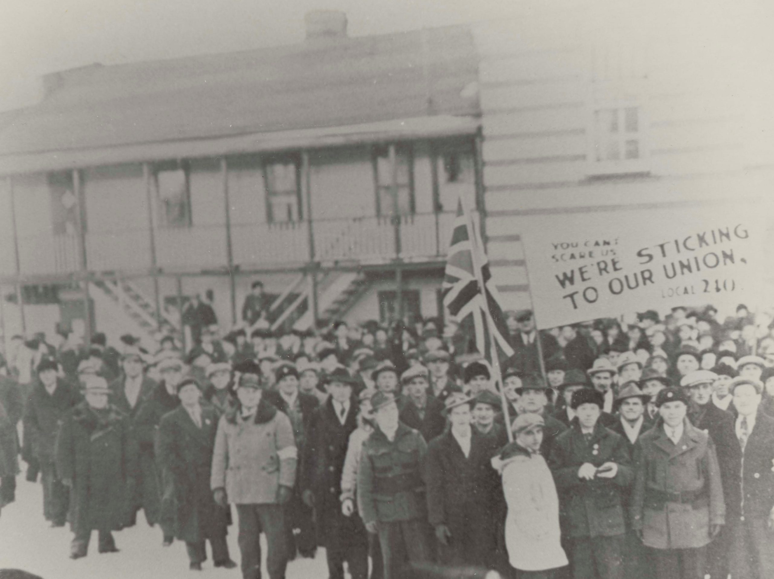 Black and white photograph of a large crowd of men dressed in warm clothing standing outside of a large building in the winter, holding a British flag and a pro-union sign.