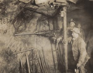 Black and white photograph of four men standing next to a large drill in a tunnel underground in a mine.