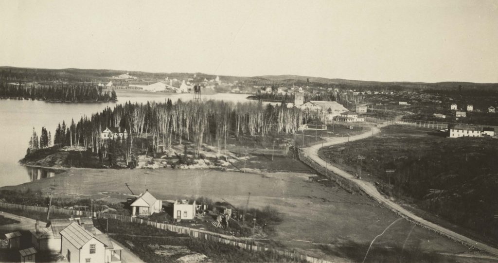 Black and white photograph taken from a high point looking down onto cleared land, mines, and houses, with a forest in the distance.