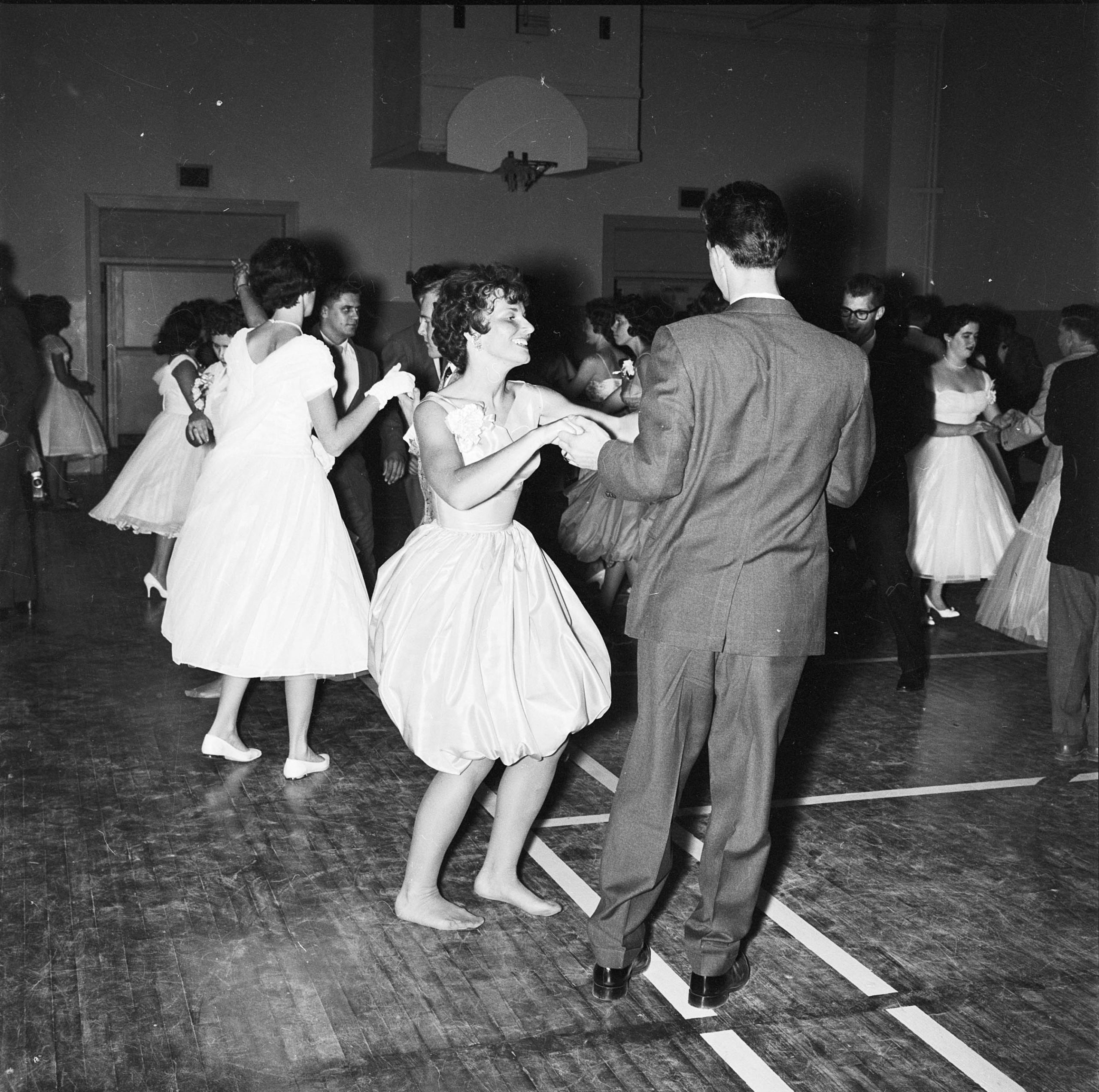 Black and white photograph of young men in suits and women in dresses dancing on a gym floor.
