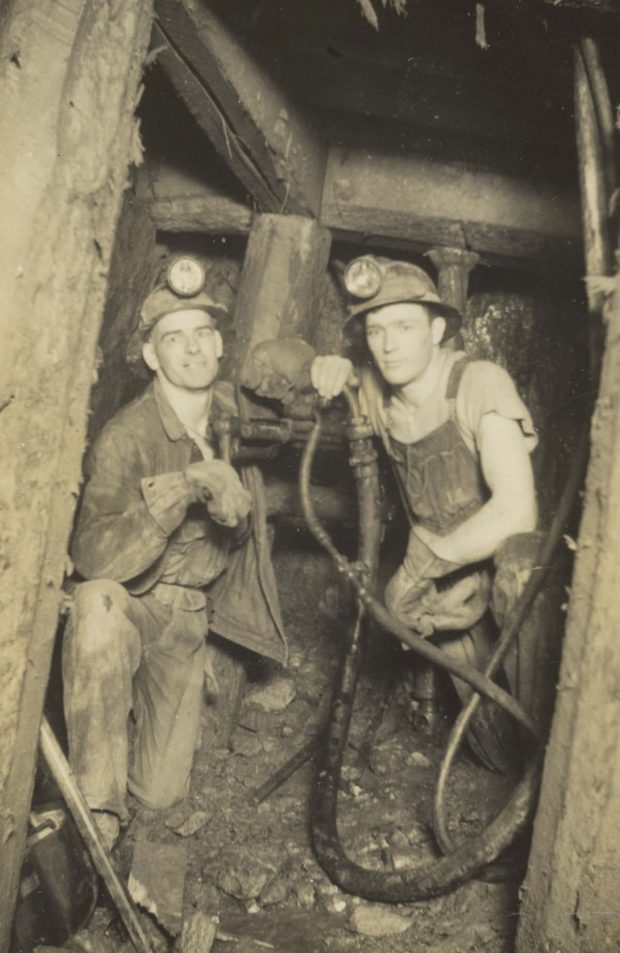 Black and white photograph of two men working underground in a mine. They are wearing hardhats with lamps and work clothes.