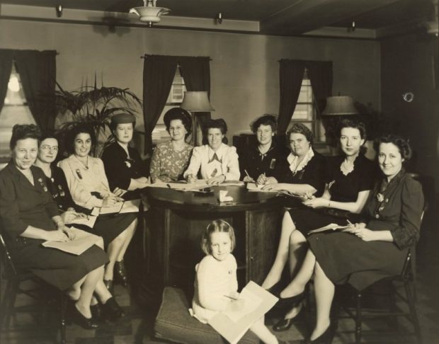 Black and white photograph of ten women seated around a large round wooden table. A girl sits on a cushion in front of the table holding a sheet of paper.