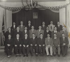 Black and white photograph of 22 men sitting and standing in front of a curtain. The men are wearing suits. A Ukrainian National Federation logo hangs on the curtain and wall behind the men.