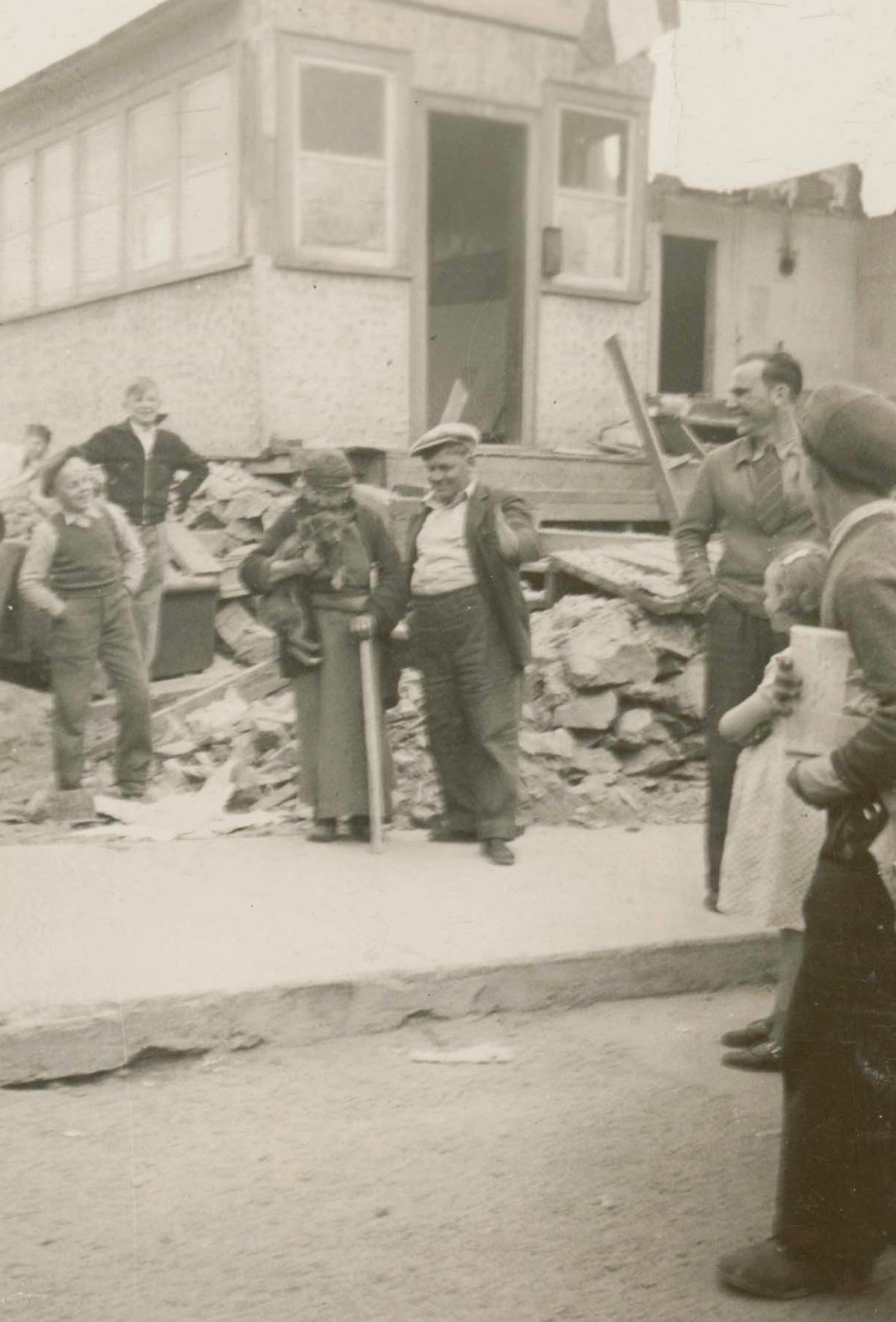 Black and white photograph taken outside of a building being demolished. A small crowd stands in front.