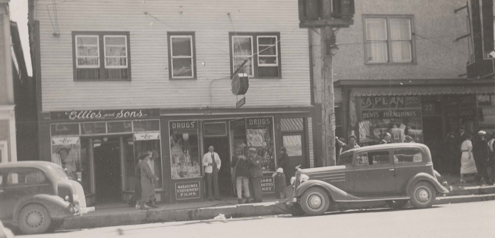 Black and white photograph of several businesses along a street. Cars are parked on the street in the foreground, with people walking on the sidewalk behind them.